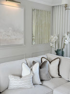 A light beige sectional sofa in a styled living room setting, decorated with several throw pillows including a white pillow with a gray geometric pattern and a textured gray pillow with a brown border.