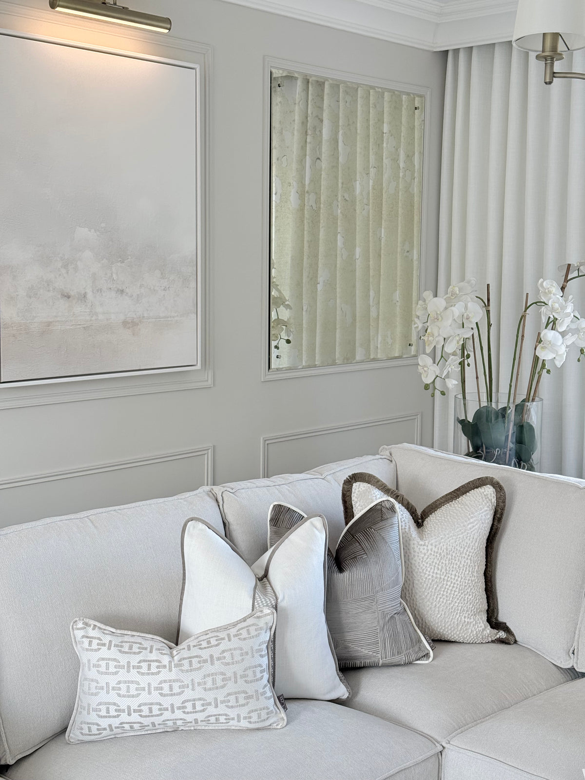 A light beige sectional sofa in a styled living room setting, decorated with several throw pillows including a white pillow with a gray geometric pattern and a textured gray pillow with a brown border.