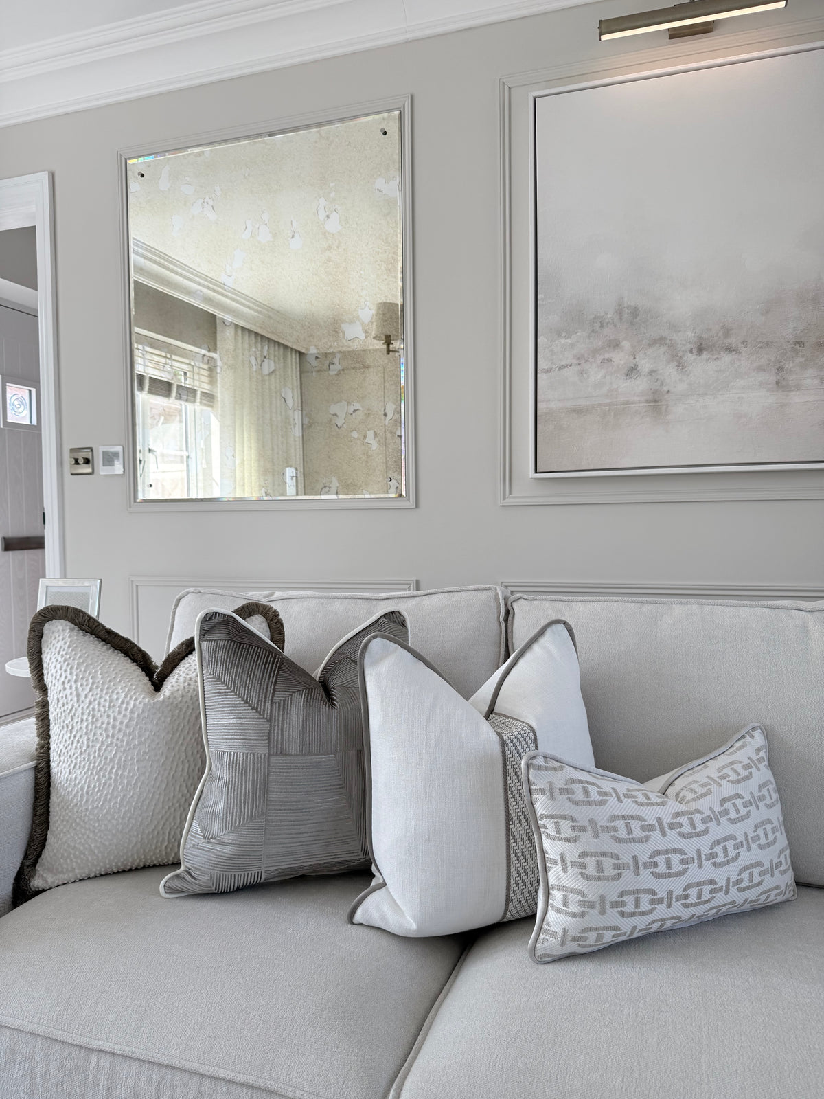 A light gray sofa in a styled living room setting, decorated with several textured and patterned throw pillows in shades of gray, white, and dark charcoal. The background features a gray paneled wall with a decorative mirror and framed abstract wall art.