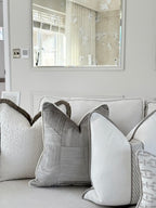 A styled collection of modern throw pillows in neutral tones, including textured cream, woven gray, and patterned beige, arranged on a light gray sofa in a bright, contemporary room.